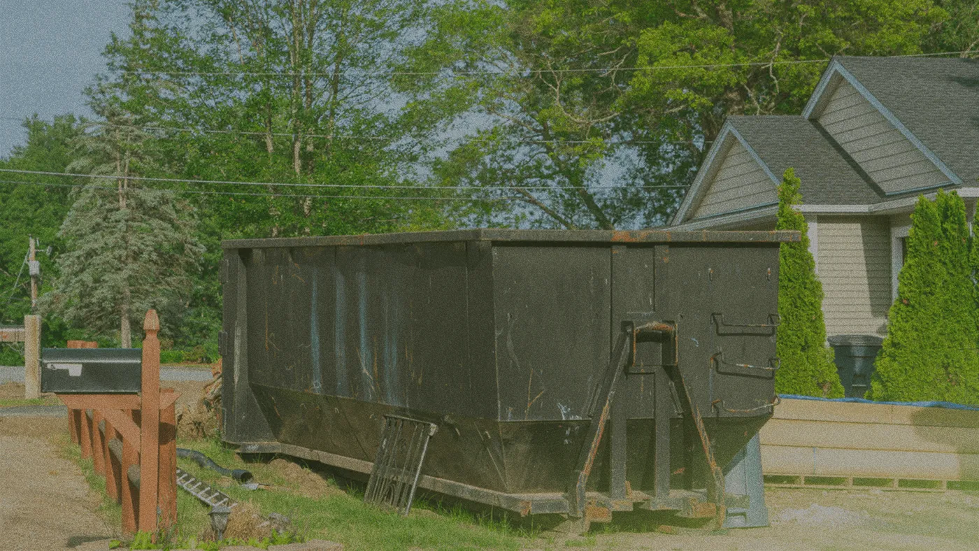 Large black dumpster on residential street, near a house with trees and bushes in the background. Construction materials nearby.