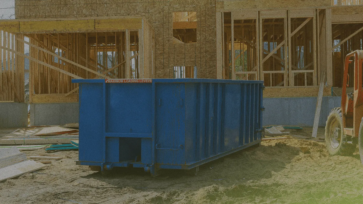 A blue dumpster sits on sandy ground at a construction site, with a partially constructed wooden building framework in the background.