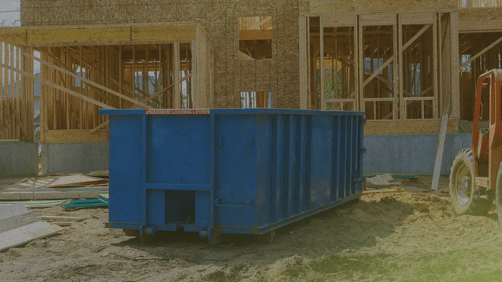 A blue dumpster sits on sandy ground at a construction site, with a partially constructed wooden building framework in the background.