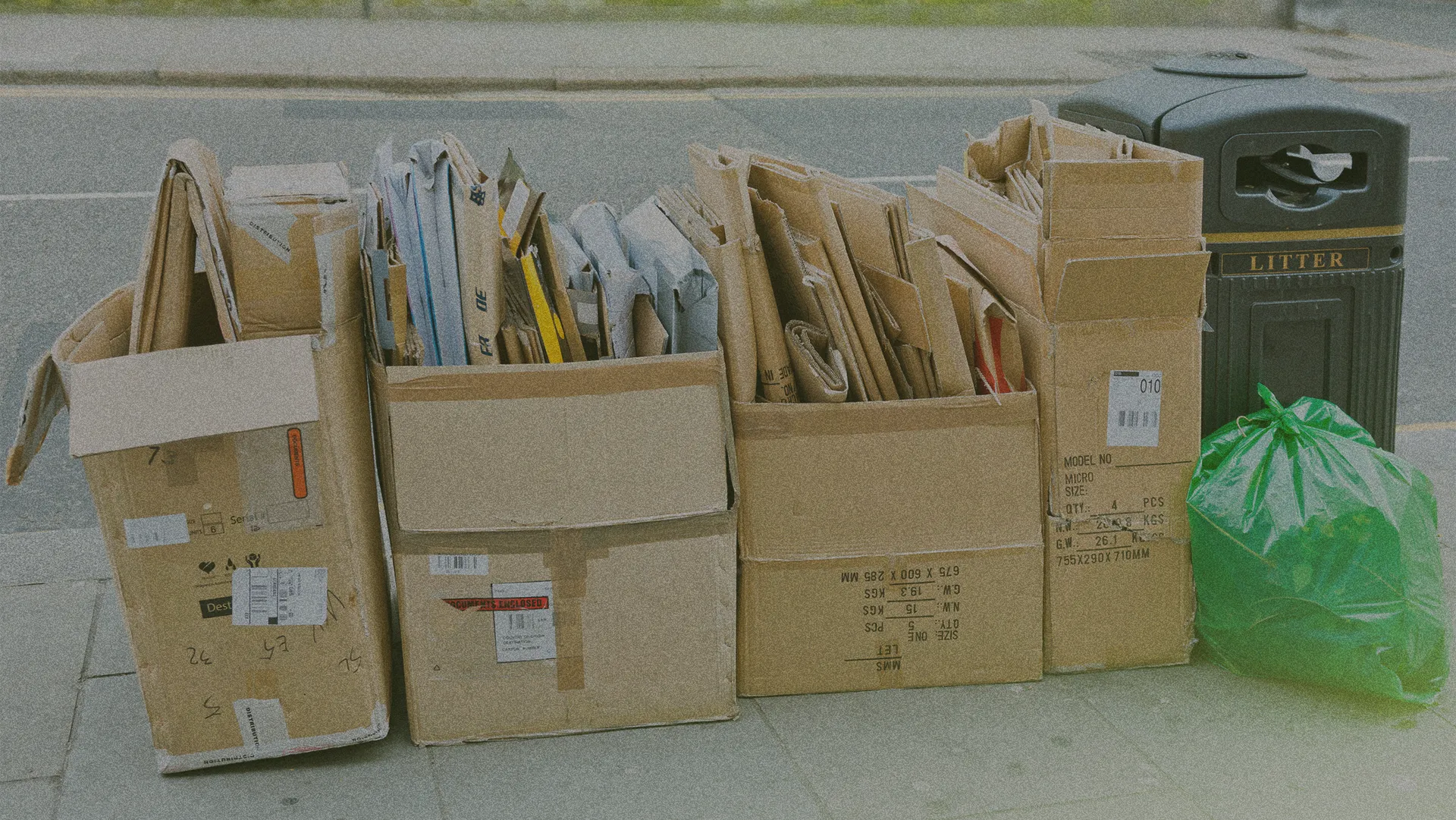 Cardboard boxes filled with flattened cartons and papers are stacked on a sidewalk next to a green garbage bag and a black litter bin.