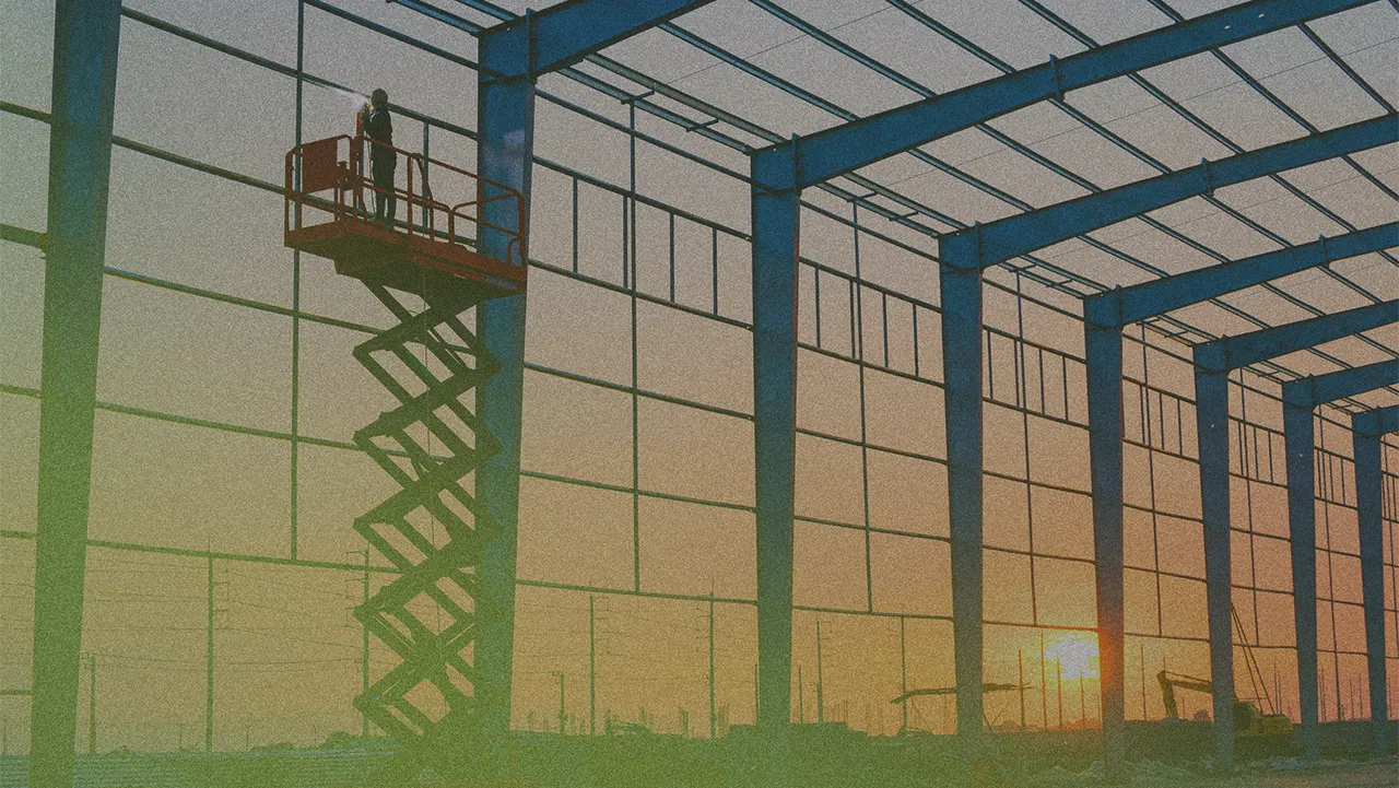 Person on a scissor lift inside a large steel structure construction site at sunset, with visible power lines and machinery in the background.