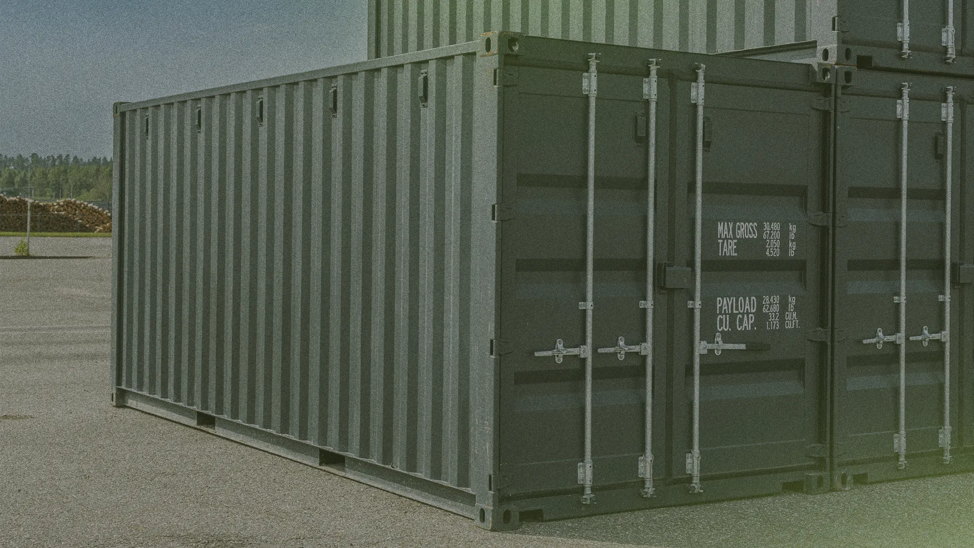 A green shipping container stacked on another, positioned on an asphalt surface in an outdoor storage area.
