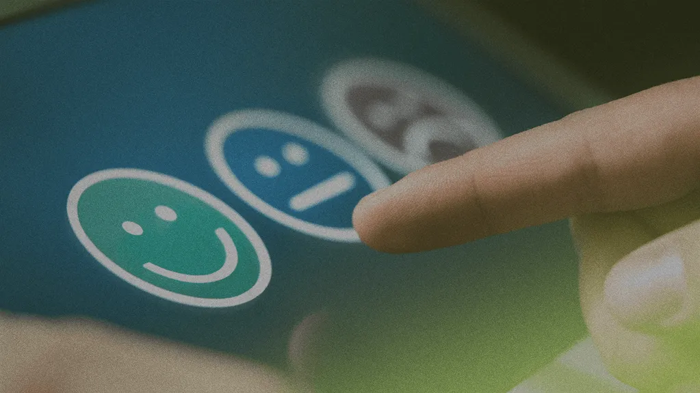 A finger pointing at a green smiling face on a touch screen, part of a feedback rating system with other face icons in the background.