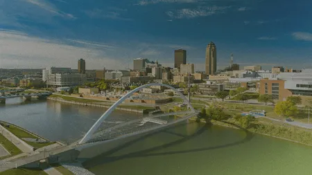 Aerial view of a cityscape with a modern arched bridge over a river, surrounded by urban buildings and greenery under a blue sky.