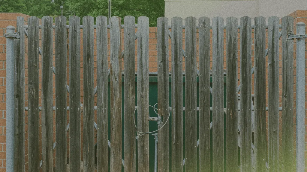 Wooden fence with vertical slats and a metal latch, set between two brick walls, with greenery visible in the background.