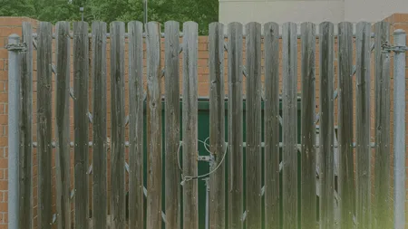 Wooden fence with vertical slats and a metal latch, set between two brick walls, with greenery visible in the background.