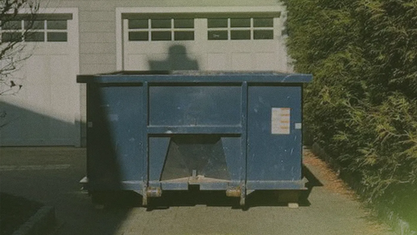 Blue dumpster in a driveway in front of closed white garage doors, a person's shadow on the door and a tall hedge to the right.