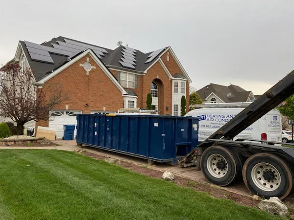A 30 yard dumpster in a driveway in front of a brick house that has solar panels