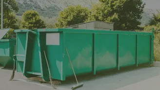 Large green dumpster on pavement with brooms leaning against it; trees and a corrugated shed in the background.