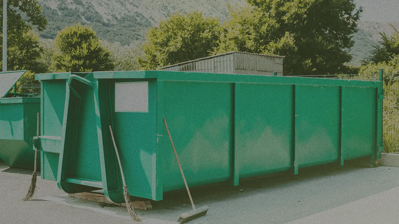 Large green dumpster on pavement with brooms leaning against it; trees and a corrugated shed in the background.