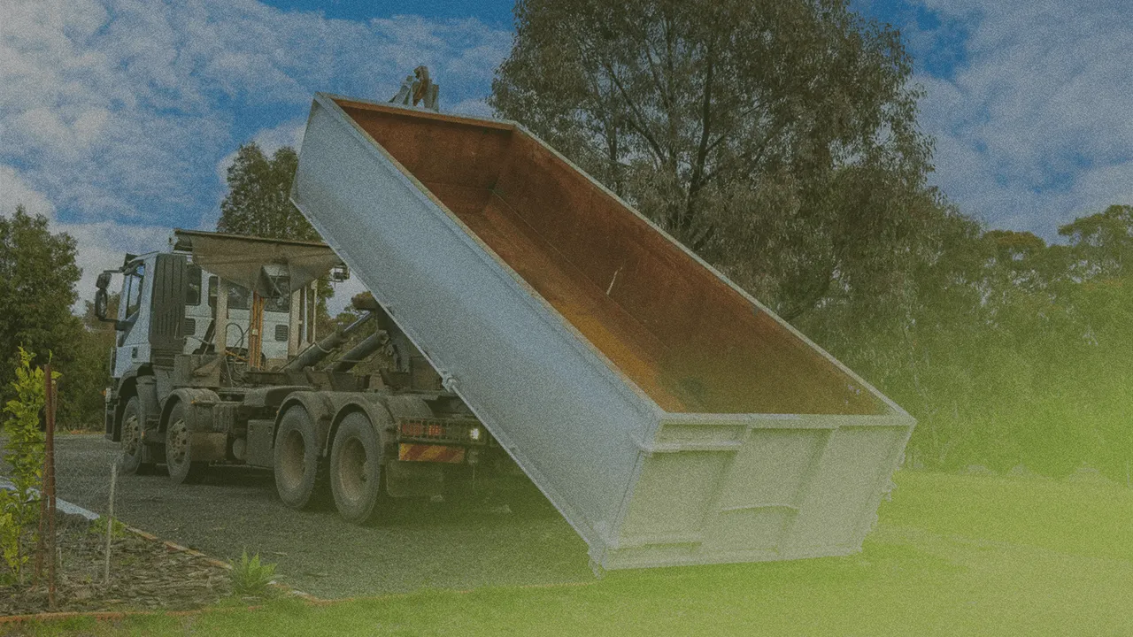 A large blue truck unloading a container onto a grassy area, surrounded by trees under a blue sky with scattered clouds.