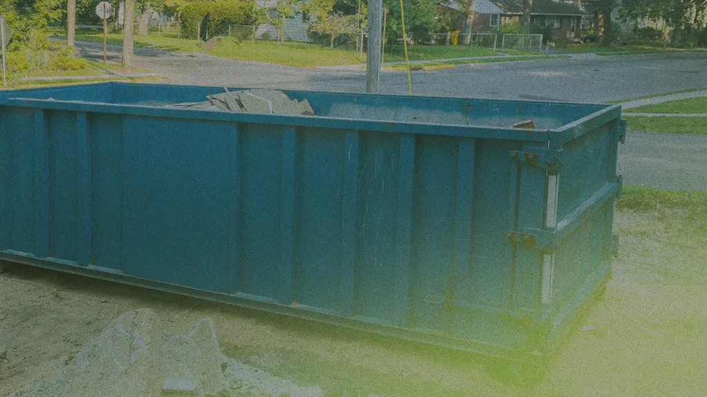 A large blue dumpster filled with debris is placed on a street, with houses and trees in the background.