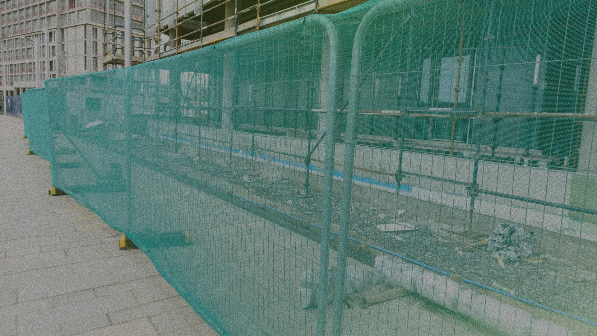A construction site with green mesh fencing along a sidewalk, showing scaffolding and building materials in the background.