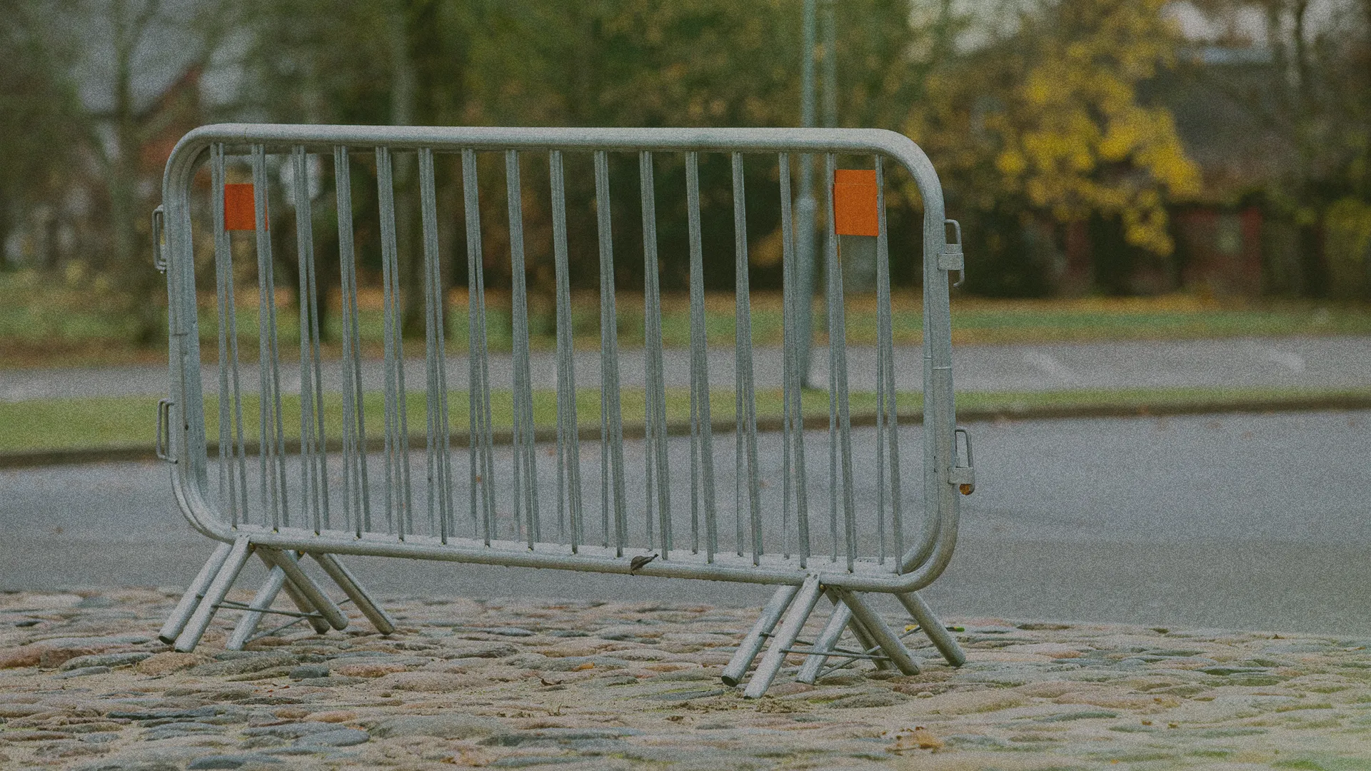 A metal barricade stands on a cobblestone path near a road, with trees and grass in the background.