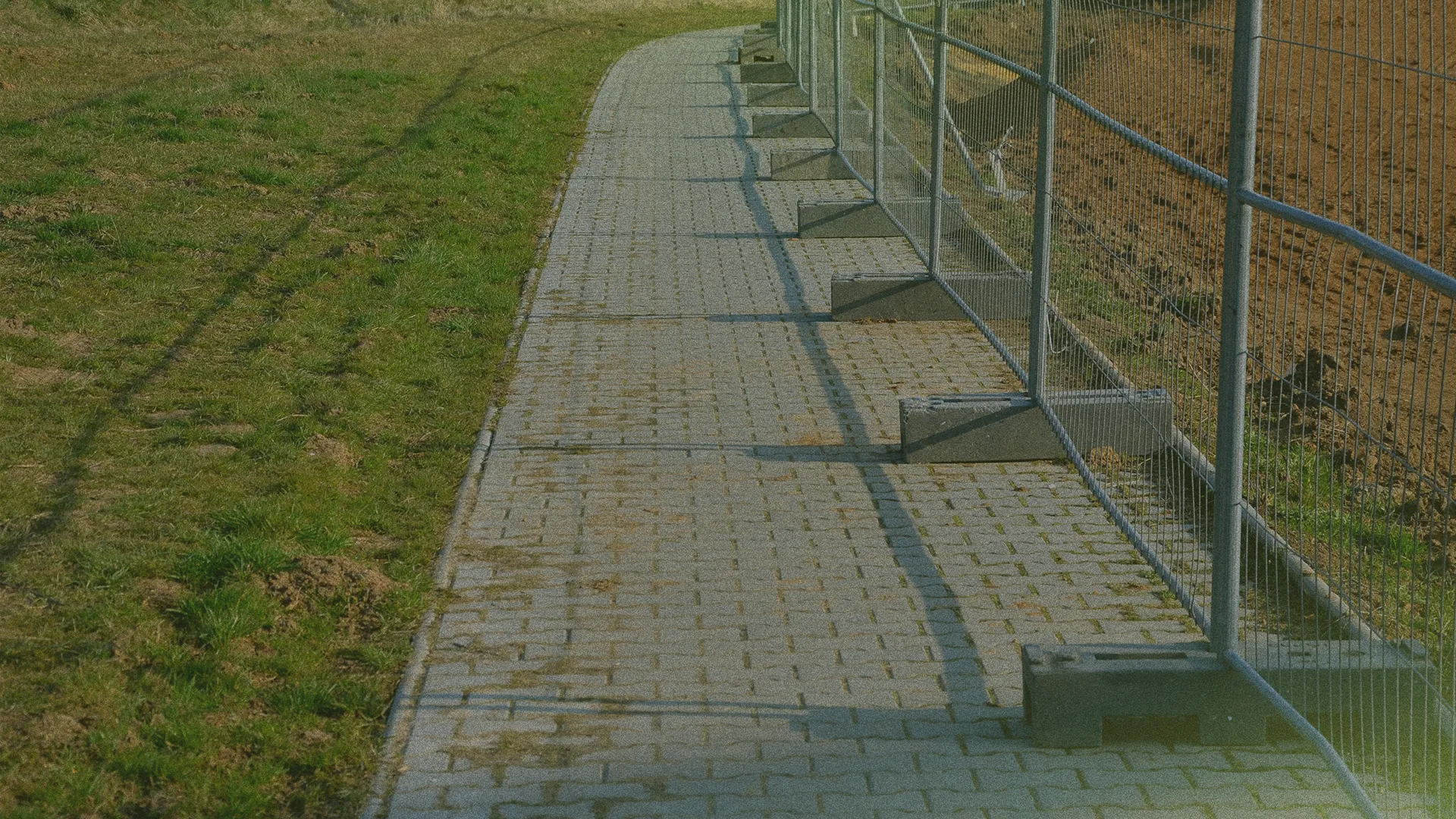 A paved walkway runs alongside a temporary metal fence, with grass on the left and soil on the right, under a clear sky.