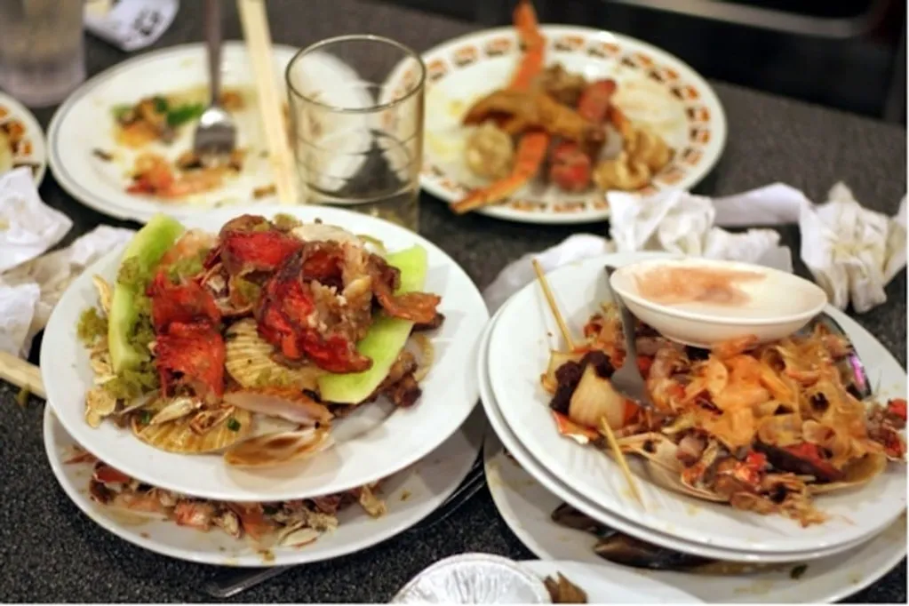 A cluttered table with plates of leftover food, crab shells, and crumpled napkins, alongside a glass and used chopsticks.