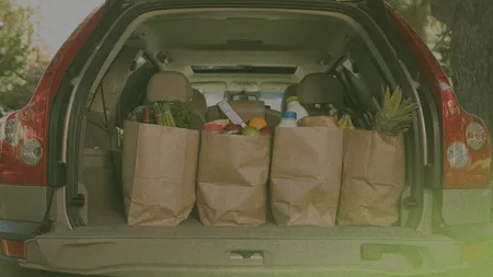 Open car trunk with four paper bags filled with groceries, including fruits, vegetables, and bread, in a parking area with trees in the background.