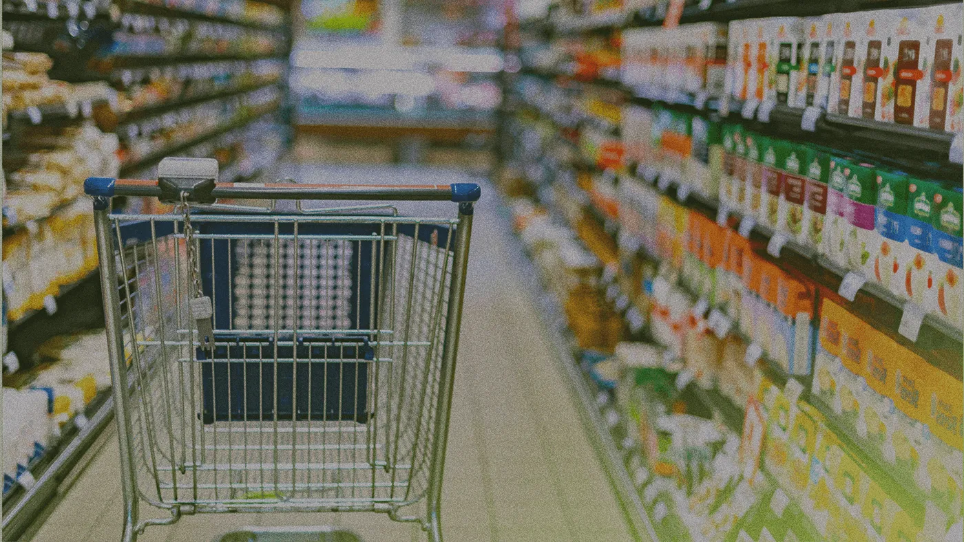 Shopping cart in a supermarket aisle, surrounded by shelves stocked with various products and colorful packaging.
