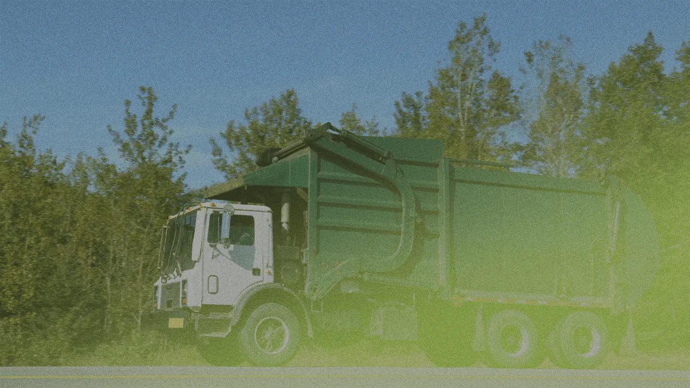 Green garbage truck driving on a road with trees in the background and bright sunlight filtering through the dust.
