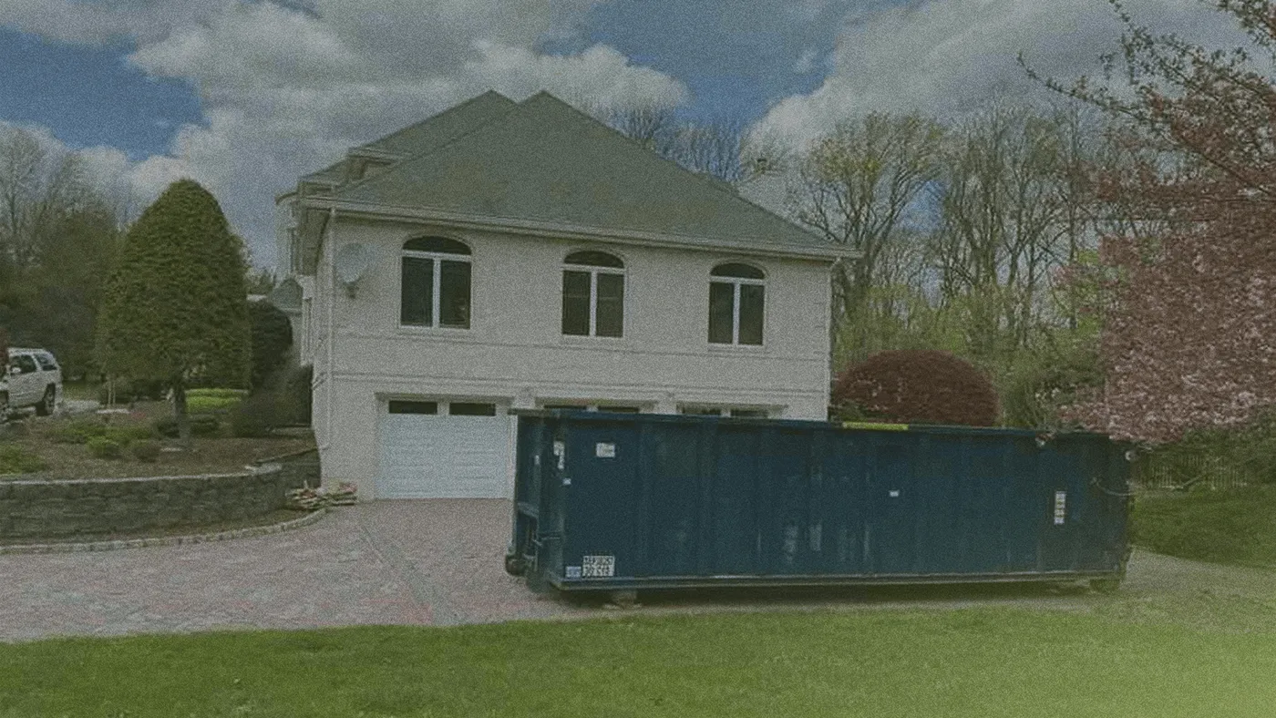 Suburban two-story house with arched upper windows and a large blue dumpster blocking the garage on a brick driveway.