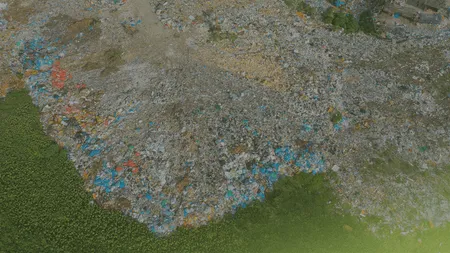 Aerial view of a large landfill with scattered trash, including blue and red plastic, adjacent to a green grassy area.