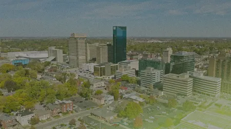 Aerial view of a cityscape with a mix of modern skyscrapers and older buildings, surrounded by lush greenery under a partly cloudy sky.