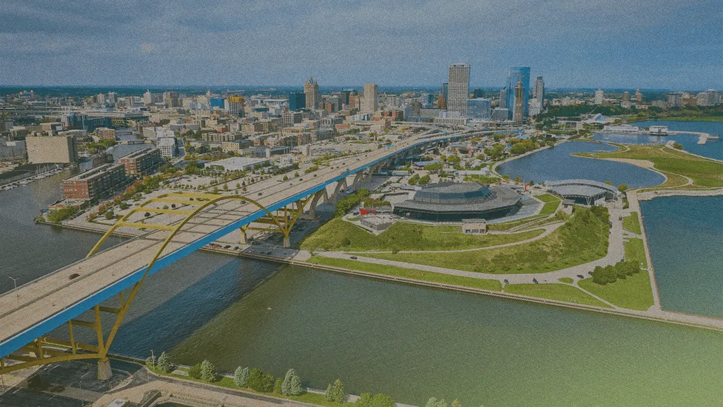 Aerial view of a cityscape with a prominent bridge, waterfront, and modern buildings under a cloudy sky.