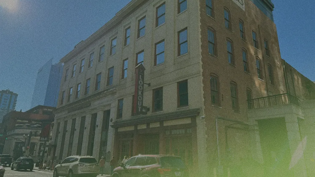 Street view of a historic three-story brick building with cars parked along the sidewalk and a sign reading "LOOKOUT" on the corner.