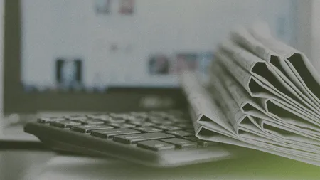 A stack of newspapers lies on a computer keyboard, with a blurred computer screen in the background.