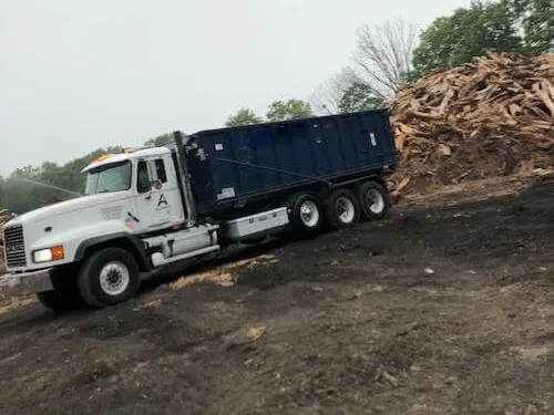 A Sourgum Waste (formerly Alliance Disposal) roll-off truck and 30 yard dumpster in front of a pile of wood