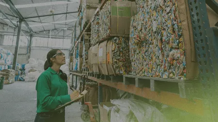 Person in a green shirt and safety goggles inspects stacked bales of recycled materials in a warehouse, holding a clipboard for notes.
