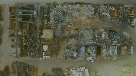 Aerial view of a scrapyard with piles of metal, wood, and various materials organized into sections, surrounded by pathways.
