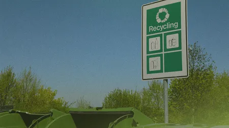 Recycling bins with open lids under a green sign showing symbols for different recyclable materials, set against a clear blue sky and trees.