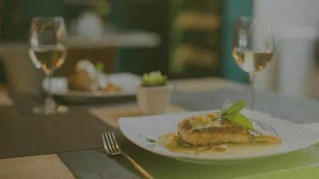 A gourmet dish with grilled fish and garnish on a white plate, set on a restaurant table with wine glasses and a small potted plant.