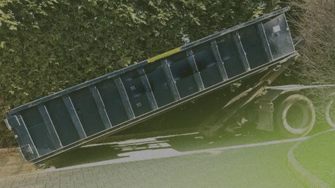 Blue roll-off dumpster tilted on a truck trailer with raised bed, parked beside a hedge on a paved surface.