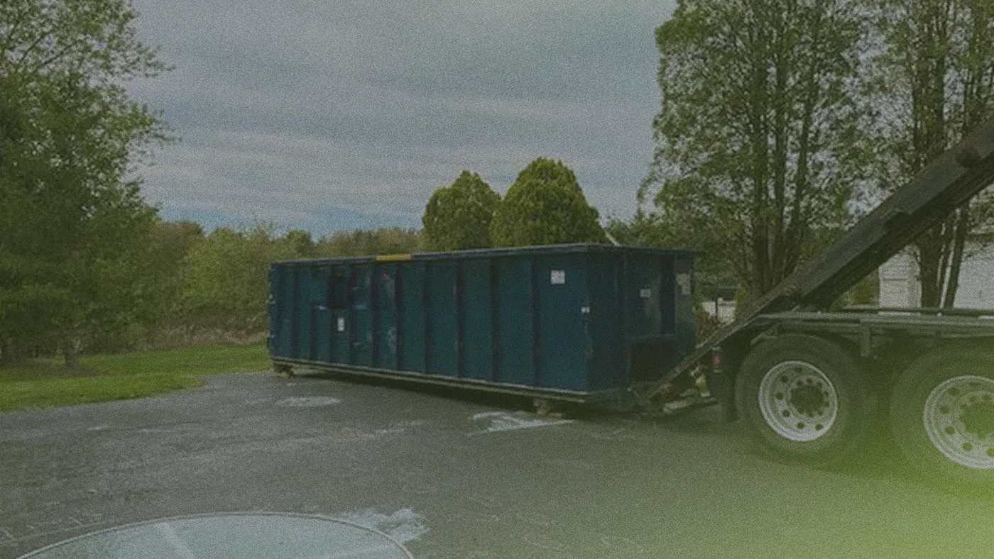 A blue dumpster container is being loaded onto a truck in a paved driveway, surrounded by trees under a cloudy sky.