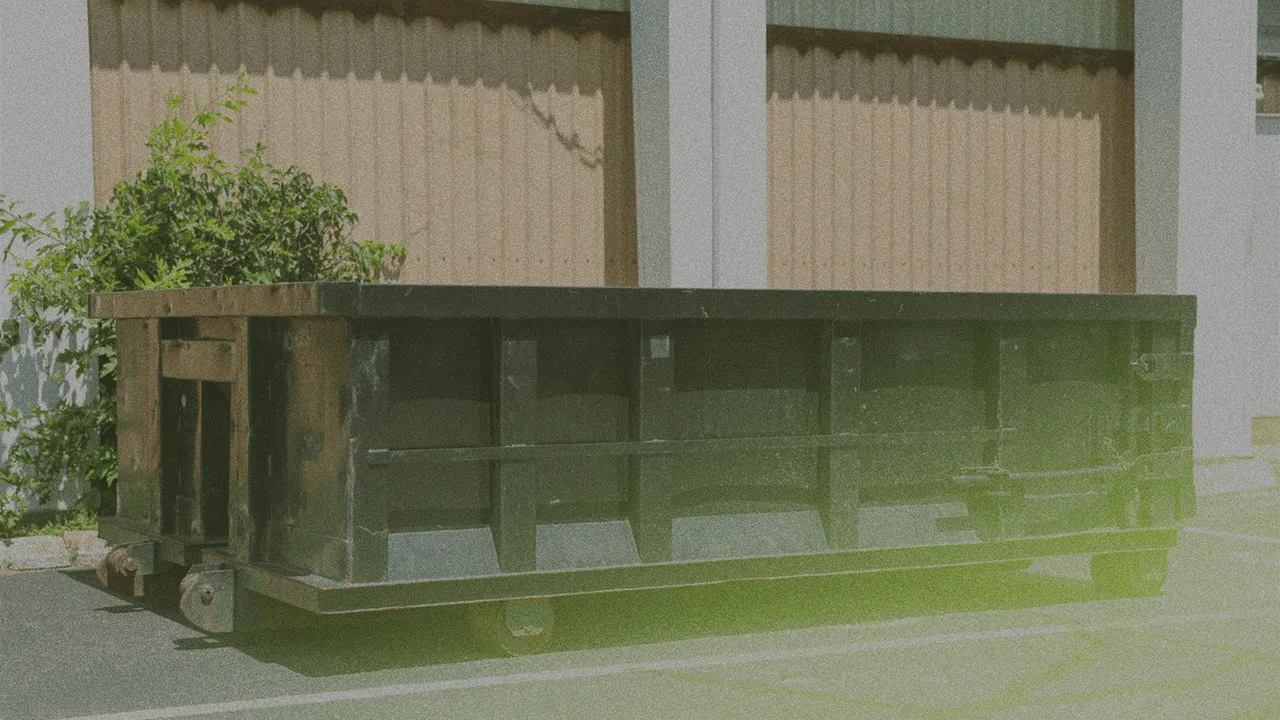 Large industrial dumpster positioned on a parking lot with green plants nearby and tall building in background.