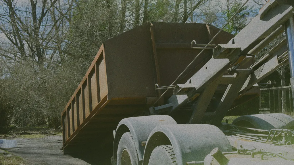 Large rusty dumpster on a truck, positioned for unloading, surrounded by trees and foliage in the background.