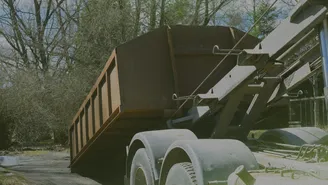 Large rusty dumpster on a truck, positioned for unloading, surrounded by trees and foliage in the background.