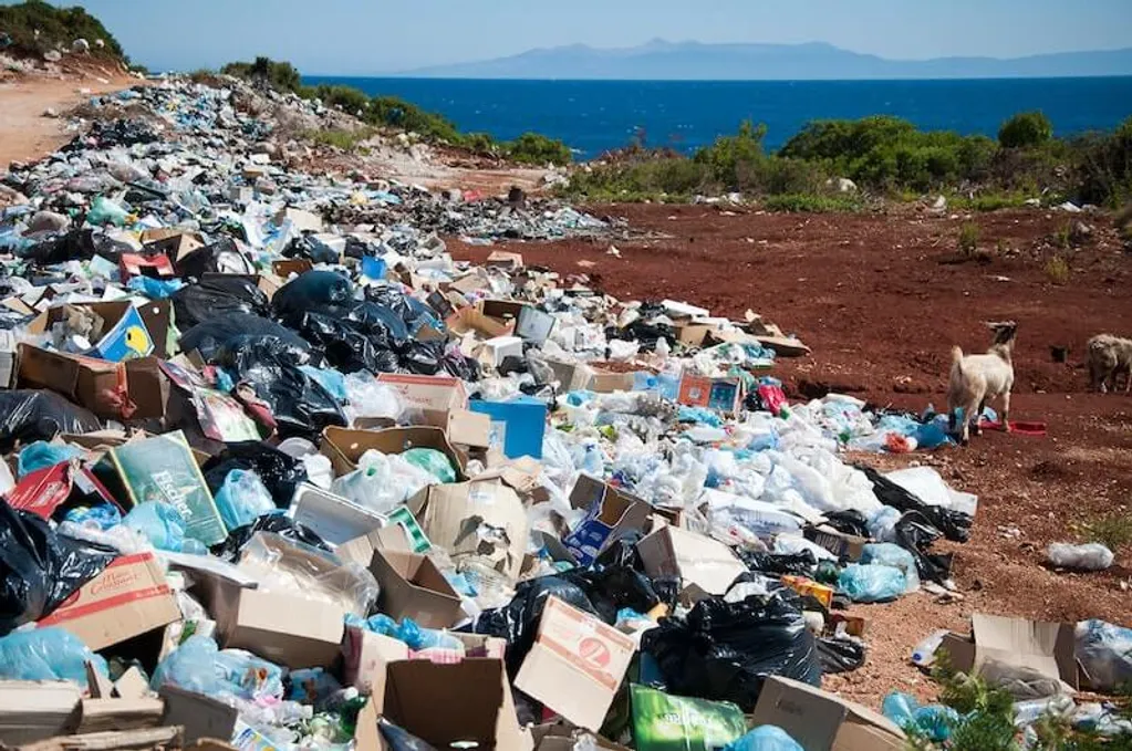 A large pile of garbage, including boxes and plastic bags, is scattered on a dirt path near the sea, with goats nearby and distant mountains visible.