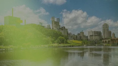 Riverfront city skyline with historic Gold Medal Flour sign, bridge and water reflections under a partly cloudy sky.