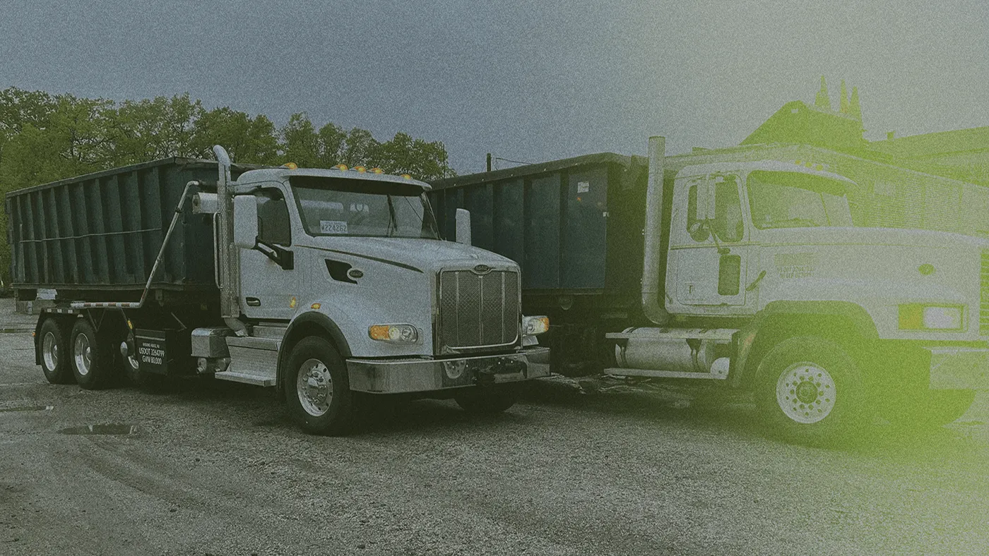 Two large dump trucks parked on a gravel surface, with trees and buildings in the background under a cloudy sky. Image has a greenish tint.