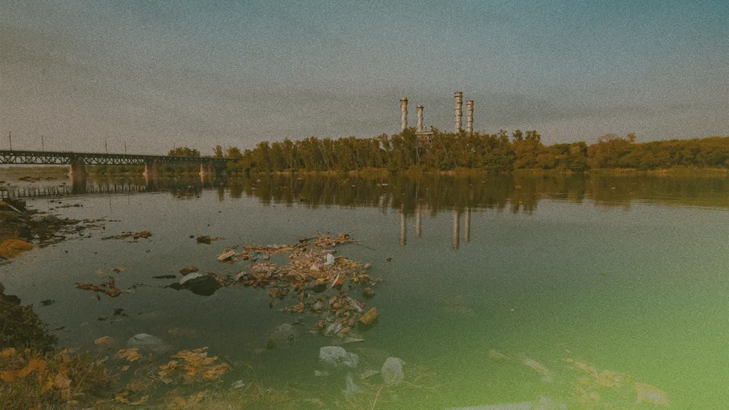 A river with floating debris, reflecting a bridge and industrial chimneys in the distance under a hazy sky.