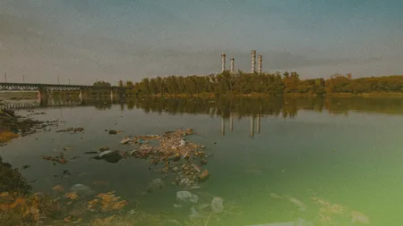 A river with floating debris, reflecting a bridge and industrial chimneys in the distance under a hazy sky.