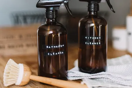 Two amber spray bottles labeled "Bathroom Cleaner" and "Kitchen Cleaner" on a wooden surface with a scrub brush and cloth nearby.
